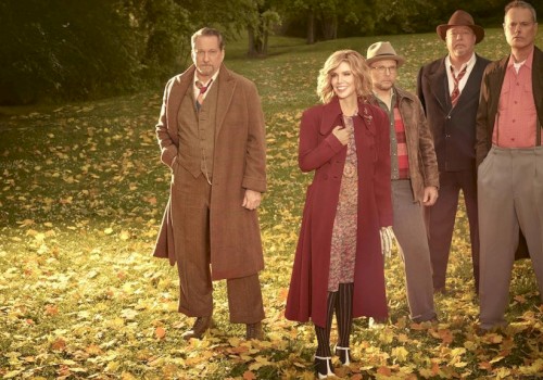 A group of five people stand outdoors in a park covered with fallen leaves, dressed in vintage, autumn-themed clothing.