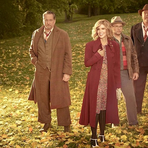 A group of five people stand outdoors in a park covered with fallen leaves, dressed in vintage, autumn-themed clothing.