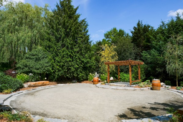 A peaceful garden scene with a wooden pergola, barrels, lush trees, and a clear blue sky in a well-maintained landscape.