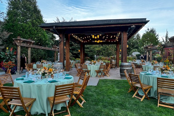 Outdoor wedding or event setup with round tables, wooden chairs, floral centerpieces, barrels, and an open pavilion under a blue sky.