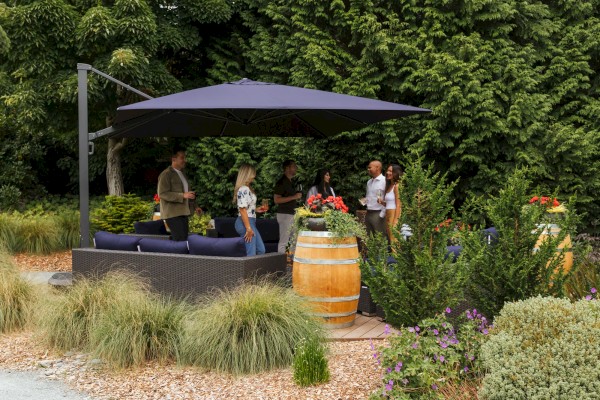 People are enjoying a garden party under a large umbrella, surrounded by lush greenery and decorative plants.