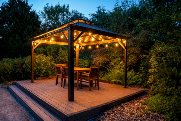 A cozy outdoor dining area with a wooden table, chairs, and warm string lights under a metal roof, surrounded by lush greenery at dusk.