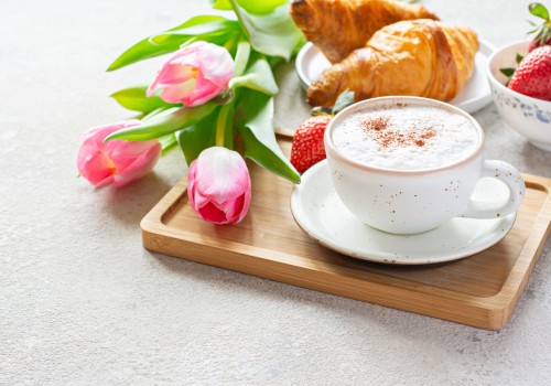 A breakfast tea with croissants, strawberries, pink tulips, and a cup of frothy coffee on a wooden tray.