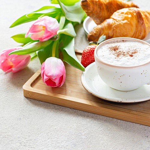A breakfast tea with croissants, strawberries, pink tulips, and a cup of frothy coffee on a wooden tray.