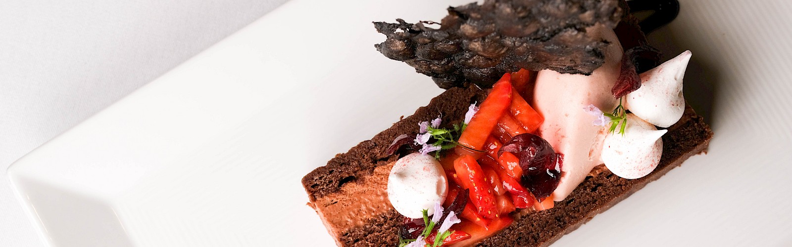 A beautifully plated chocolate dessert with strawberries, whipped cream, edible flowers, and a decorative chocolate wafer on a white plate.