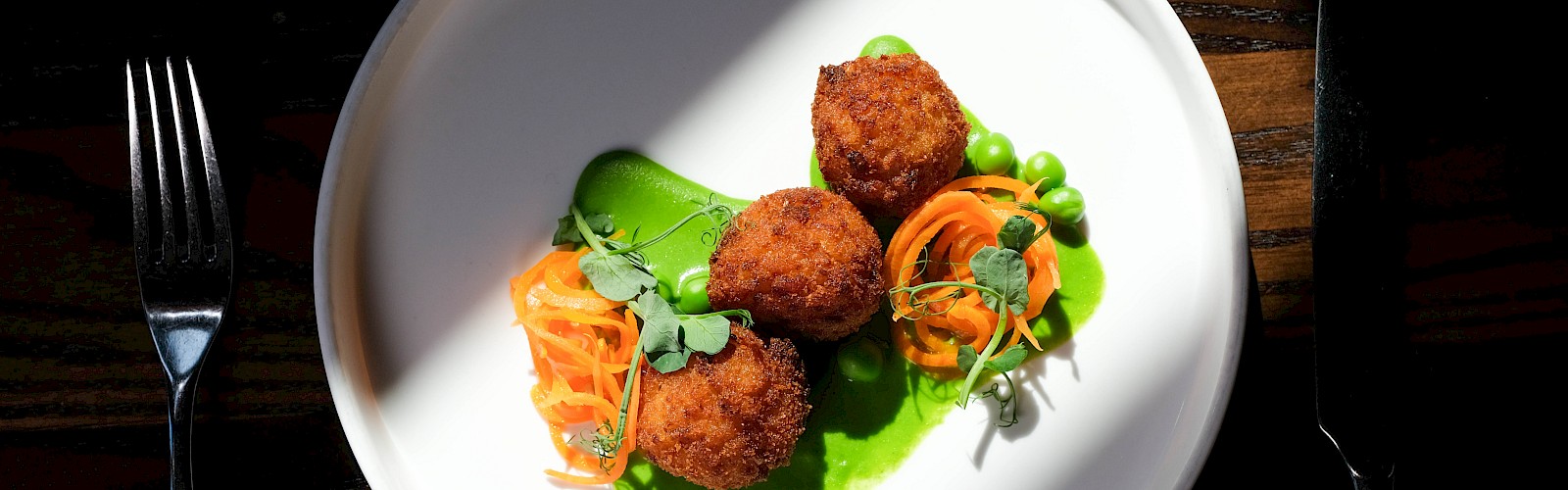 A dish with fried croquettes, carrots, peas, and green sauce on a white plate, placed on a dark wooden table.