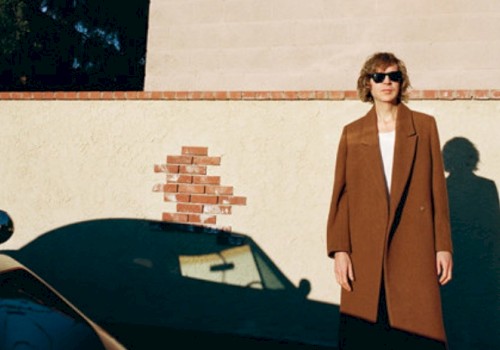 A woman with curly hair wearing sunglasses and a brown coat stands against a beige wall with a brick patch, shadow cast on the wall.