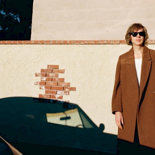A woman with curly hair wearing sunglasses and a brown coat stands against a beige wall with a brick patch, shadow cast on the wall.