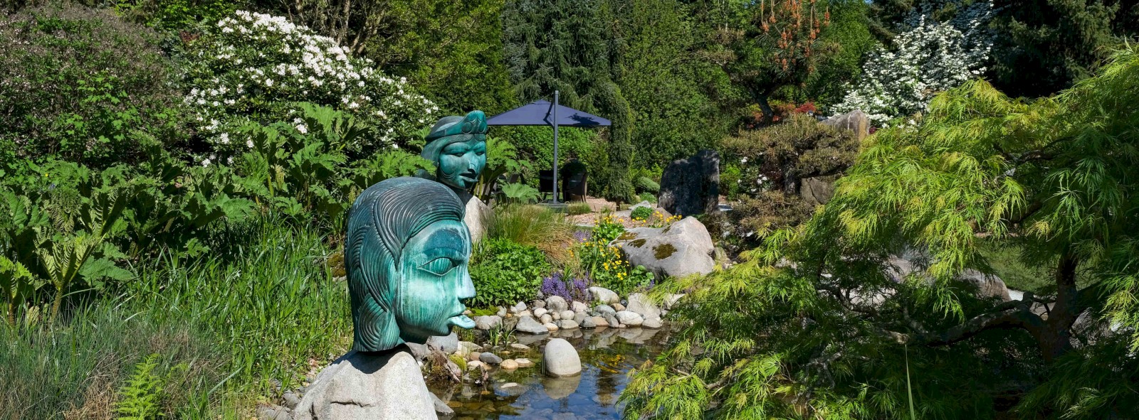 A lush garden scene with ceramic head sculptures near a small pond, surrounded by green plants, trees, rocks, and colorful flowers.