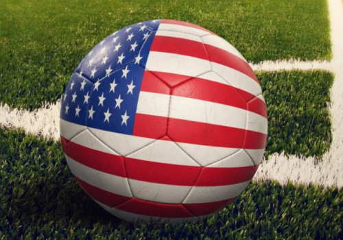 A soccer ball with an American flag design is on a green field with white lines, likely on a stadium or sports ground.