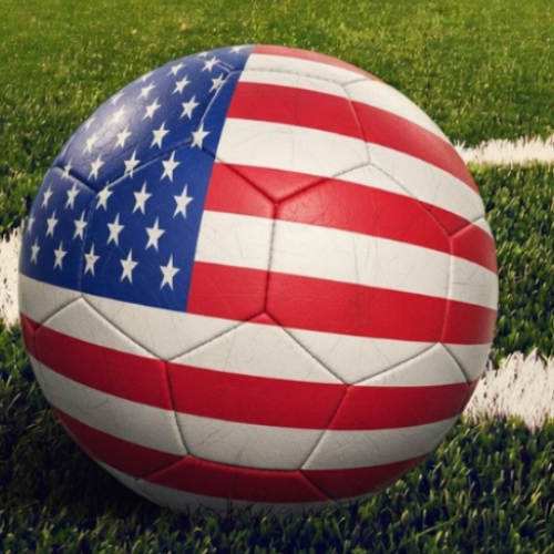 A soccer ball with an American flag design is on a green field with white lines, likely on a stadium or sports ground.