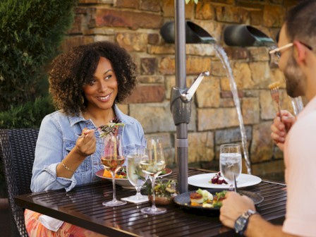 A couple enjoys a romantic outdoor dinner with wine and desserts near a brick wall and water feature.