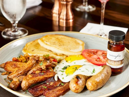 A hearty breakfast plate with eggs, bacon, sausage, pancakes, tomato, and syrup, accompanied by glasses of water and wine on a wood table.