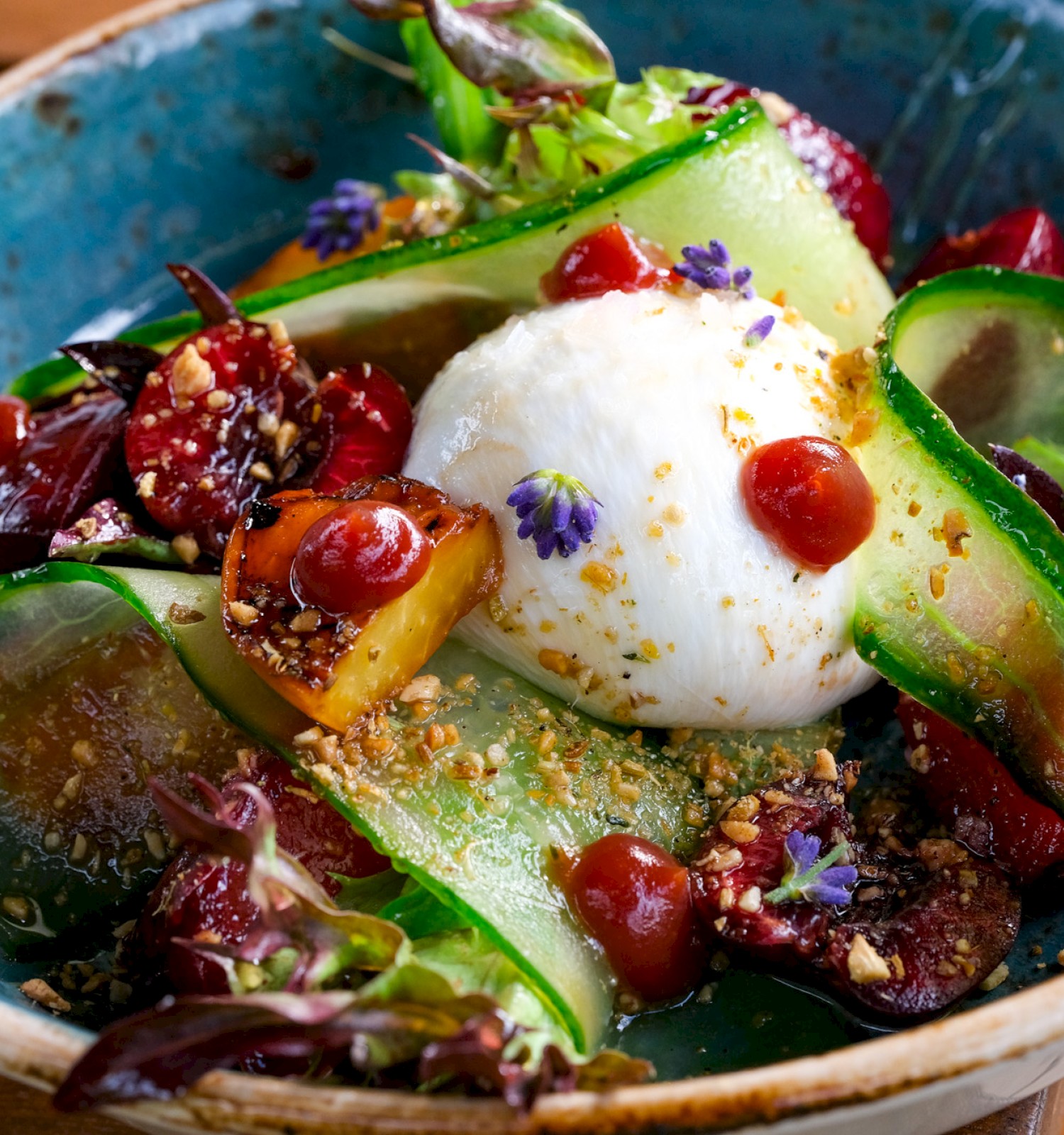 A vibrant salad with cucumber, cherry tomatoes, roasted vegetables, a poached egg, edible flowers, and sprinkled nuts in a rustic bowl.