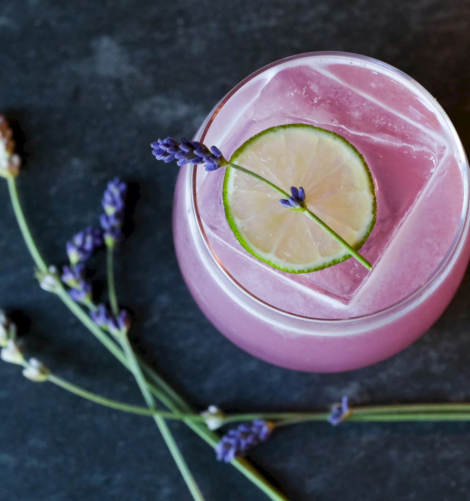 A pink drink with a lemon slice and lavender garnish in a glass on a dark surface, plus loose lavender sprigs beside it.