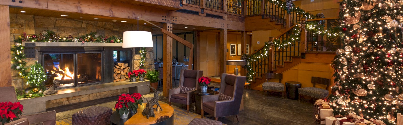 A cozy, festive living room decorated for Christmas with a large tree, wrapped presents, a fireplace, and holiday ornaments.