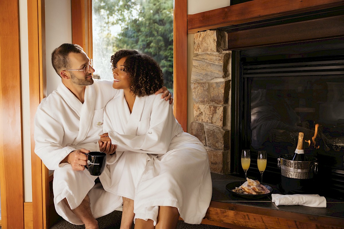 A couple in white robes sharing a tender moment by a cozy fireplace with champagne and snacks, enjoying a relaxed morning together.
