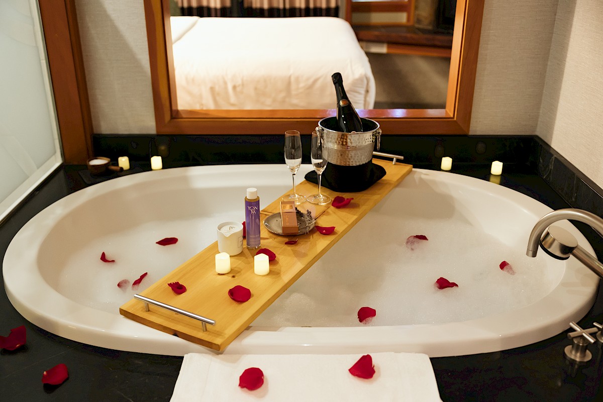 A romantic bubble bath setup with rose petals, candles, champagne, and toiletries in a hotel bathroom.