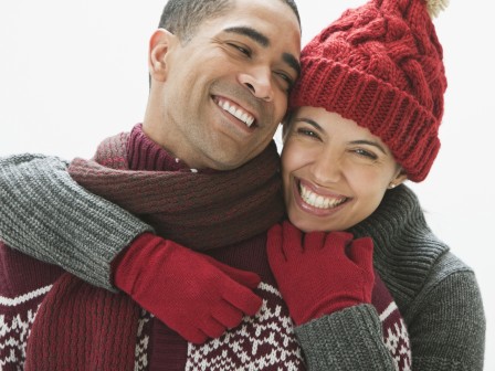 A smiling couple dressed in winter clothes, embracing each other, with both wearing red gloves and a red knit hat.