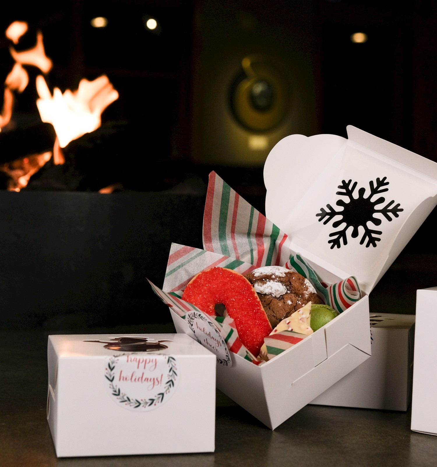 A box of assorted holiday cookies inside a festive box, with Christmas decorations and a glowing fire in the background.