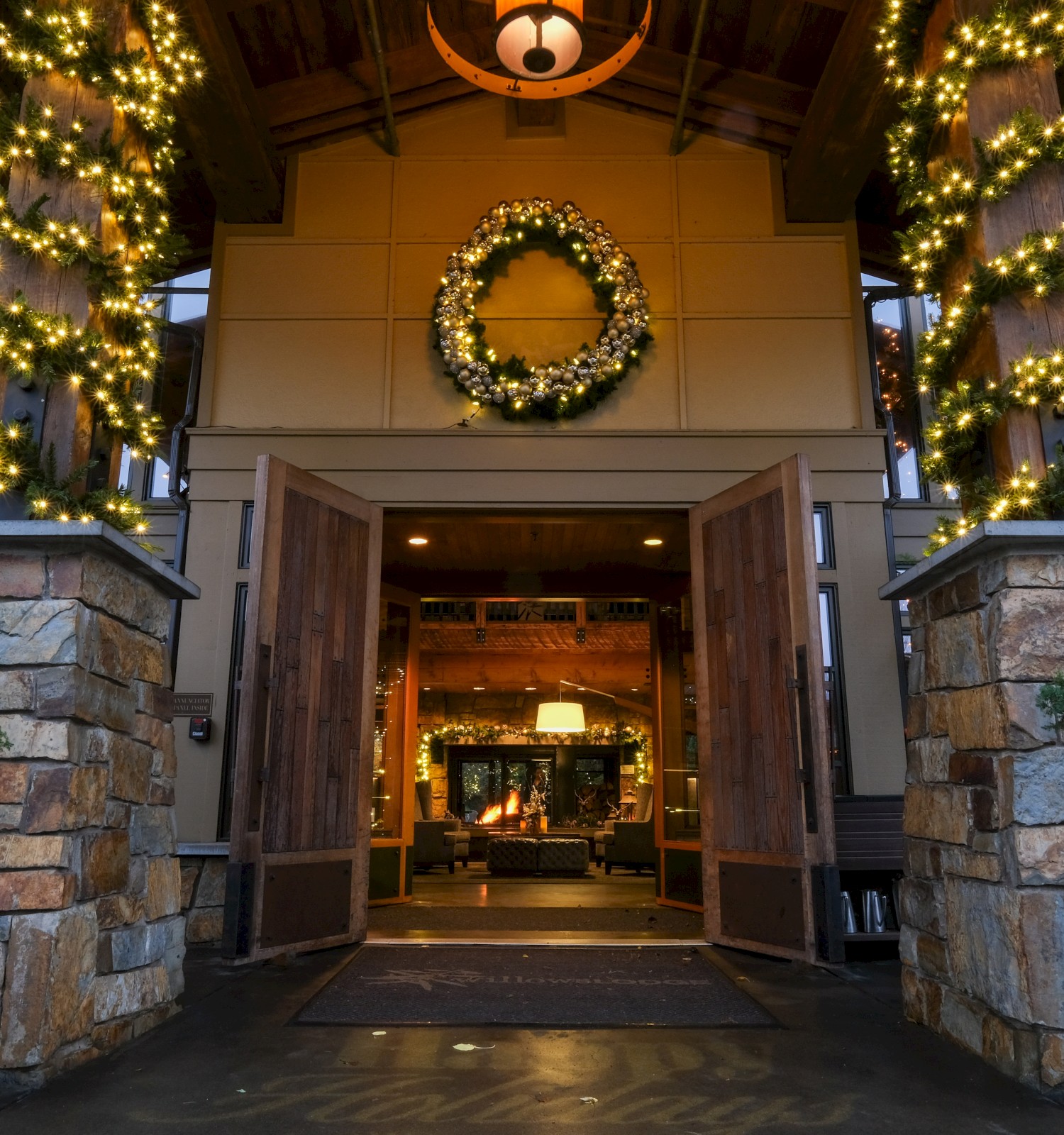A warmly decorated house entrance with Christmas lights, a wreath, and a cozy interior visible through open wooden doors.