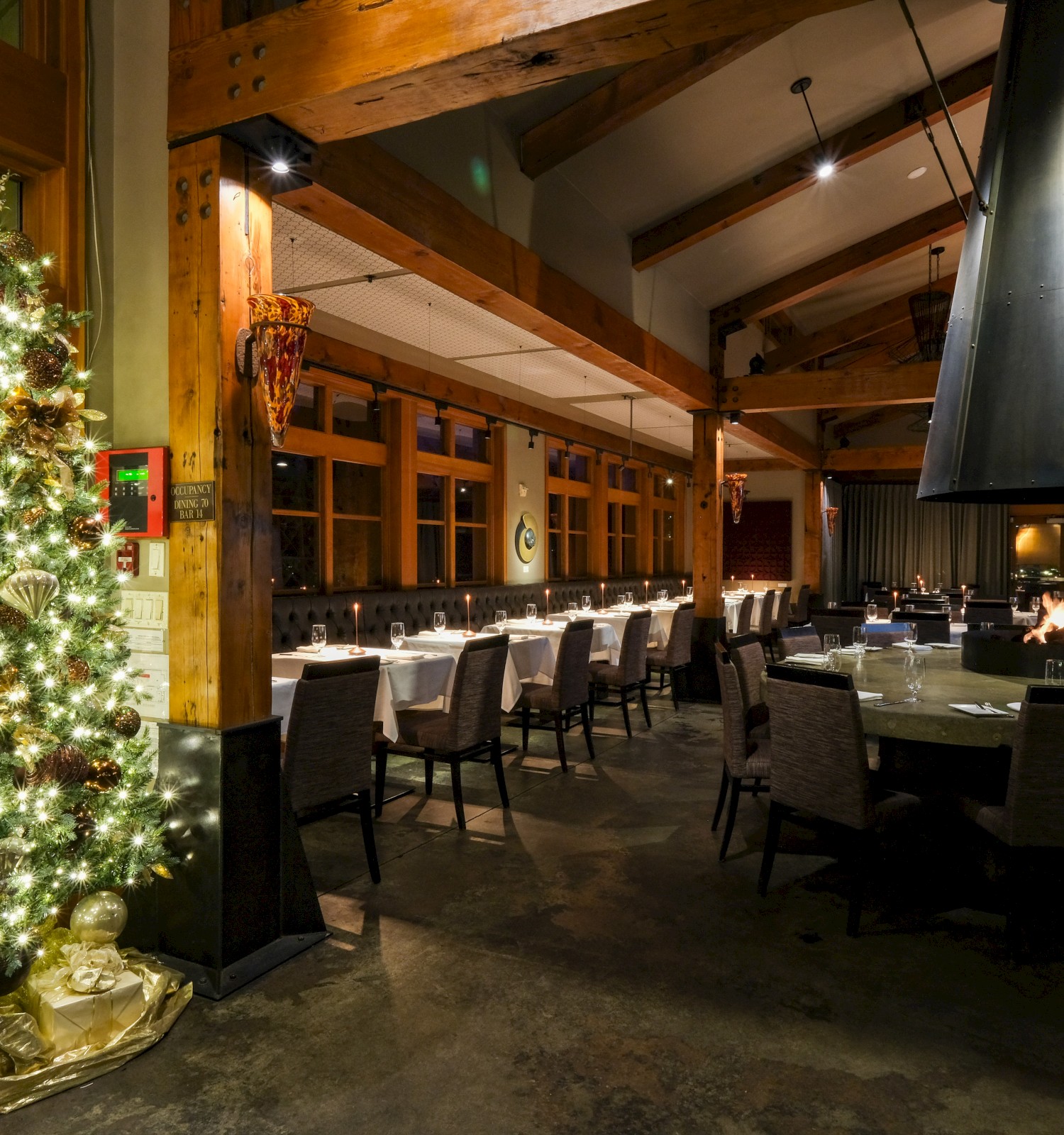 A warmly lit, festive restaurant interior decorated for Christmas with a Christmas tree, tables set for dining, and a central fireplace.