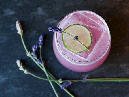 A pink drink with a lemon slice and lavender, garnished with a lavender sprig, on a dark surface next to some lavender flowers.