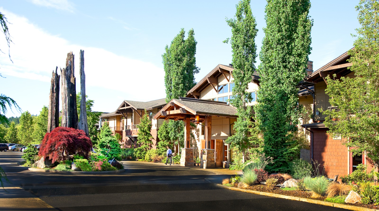 A picturesque lodge with tall green trees, a wooden entrance, and lush landscaping under a clear blue sky, showcasing a serene and inviting environment.