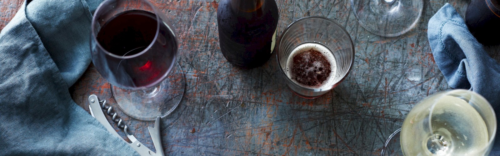 Various glasses of wine and beer, a bottle, a corkscrew, and napkins are on a rustic table.