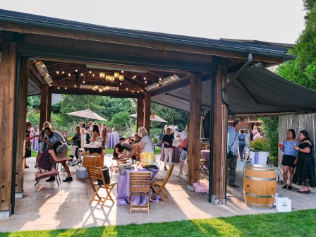 People gather at an outdoor event under a wooden pavilion with string lights, enjoying food, drinks, and socializing.