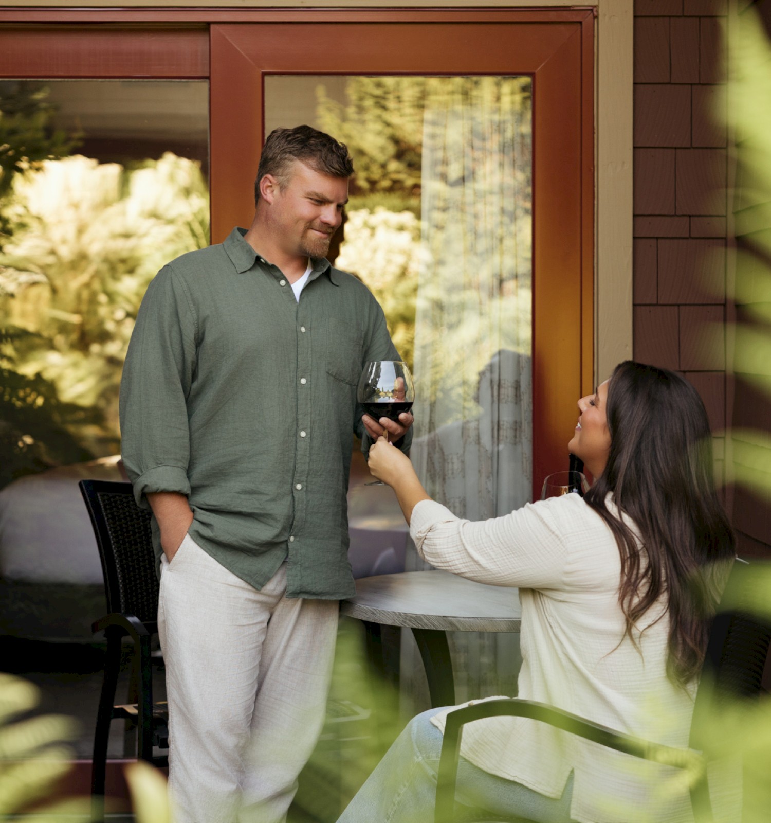 A woman offers a glass of red wine to a man on a patio, both smiling, in a cozy outdoor setting.