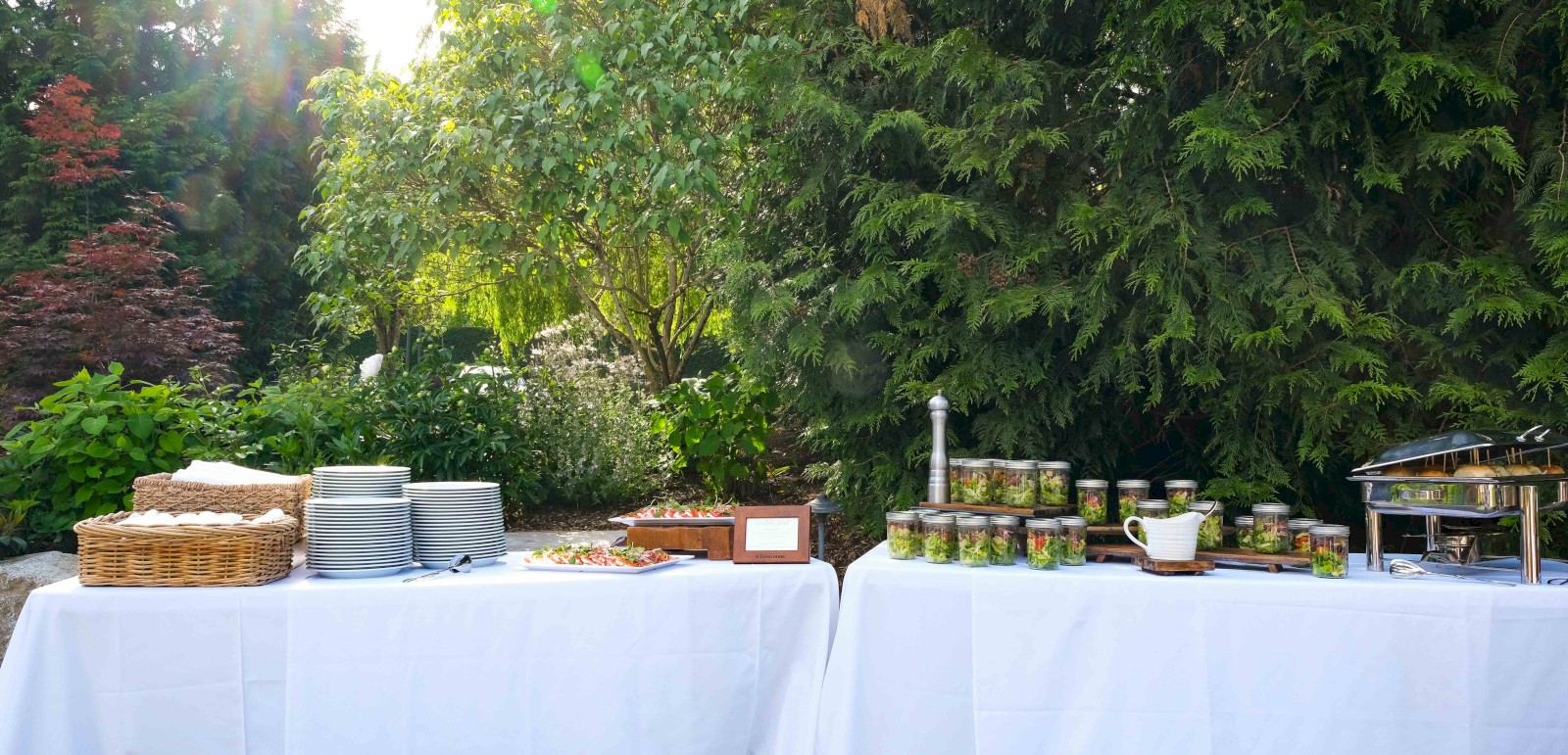 This is an outdoor buffet setup with plates, salads in jars, a chafing dish, and cutlery, set against lush green trees and sunlight.