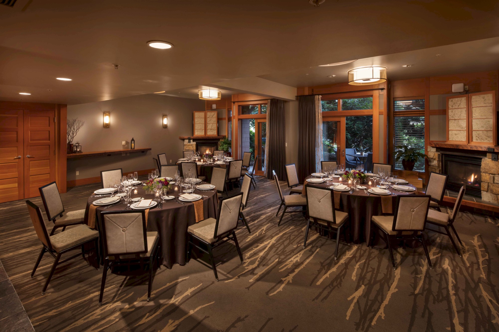 A warmly lit dining room with round tables set for a meal, upholstered chairs, and large windows showing greenery outside.