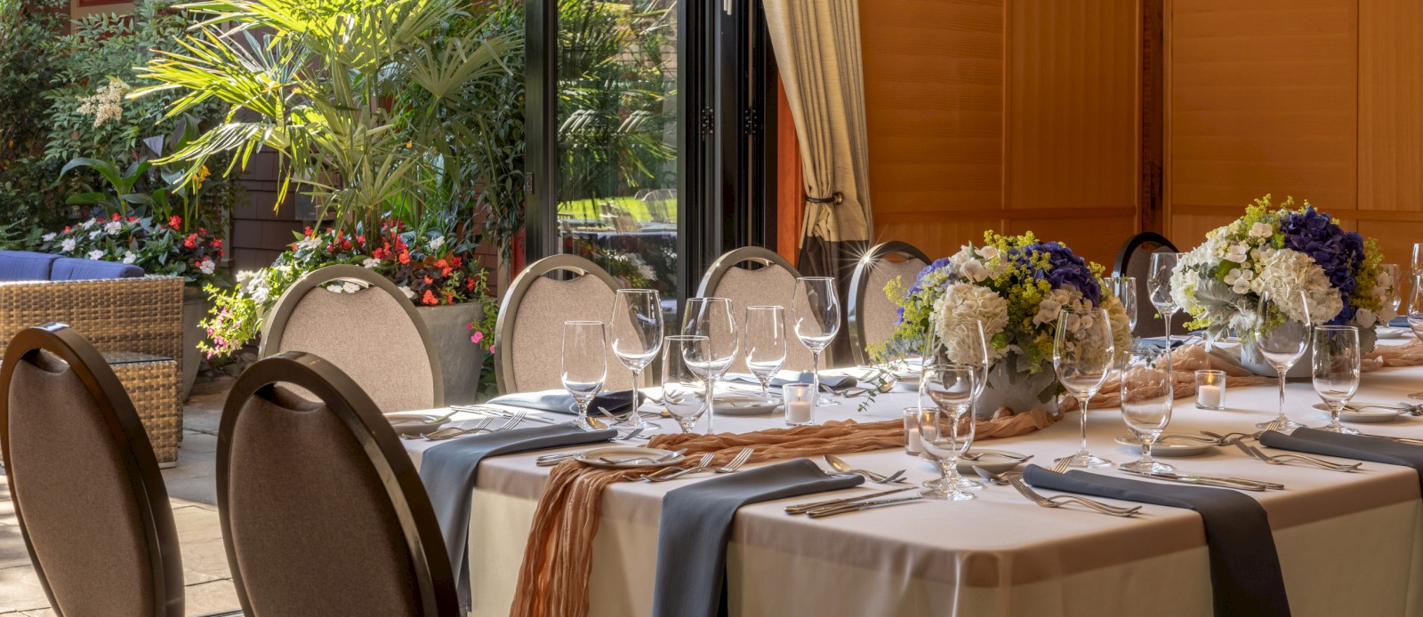 A beautifully set dining table with white tablecloth, floral centerpieces, and glassware inside a room with an open view of lush outdoor greenery.