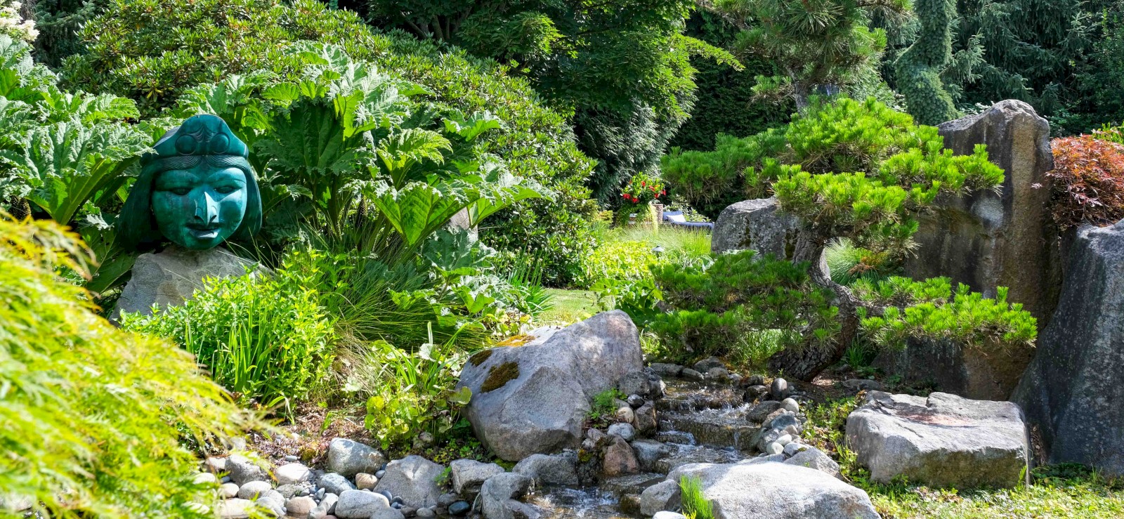 A lush garden scene features rocks, a small stream, varied green plants, and a sculpture with a blue face partially hidden among the foliage.
