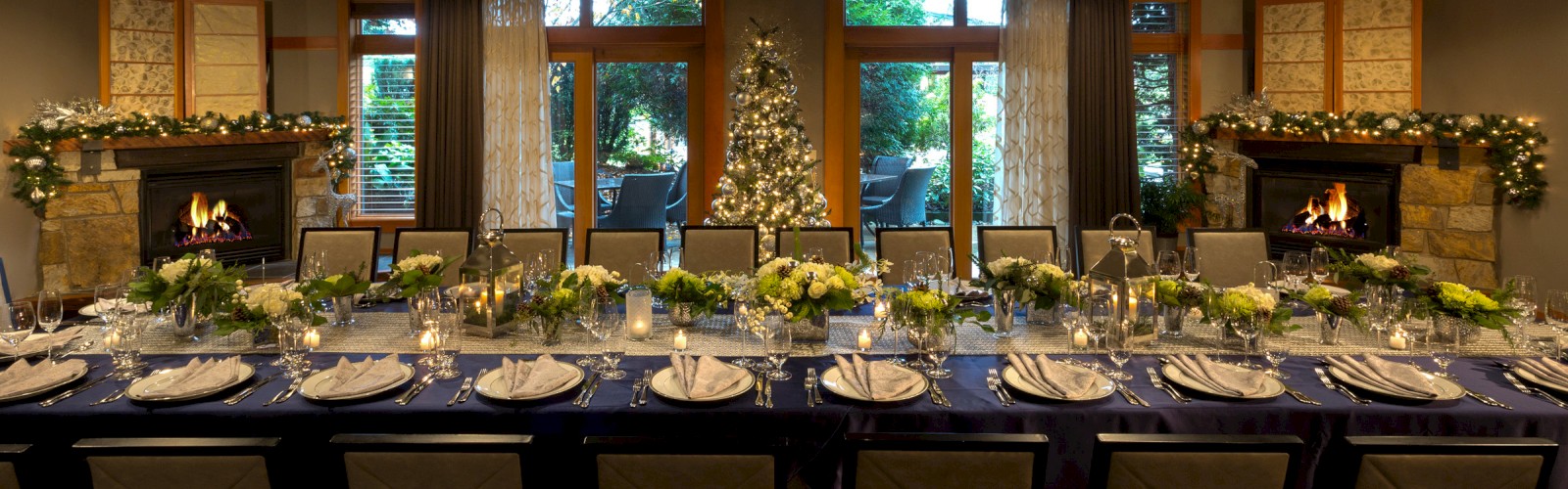 A beautifully decorated holiday dinner table with candles, floral arrangements, and a Christmas tree in front of large windows.
