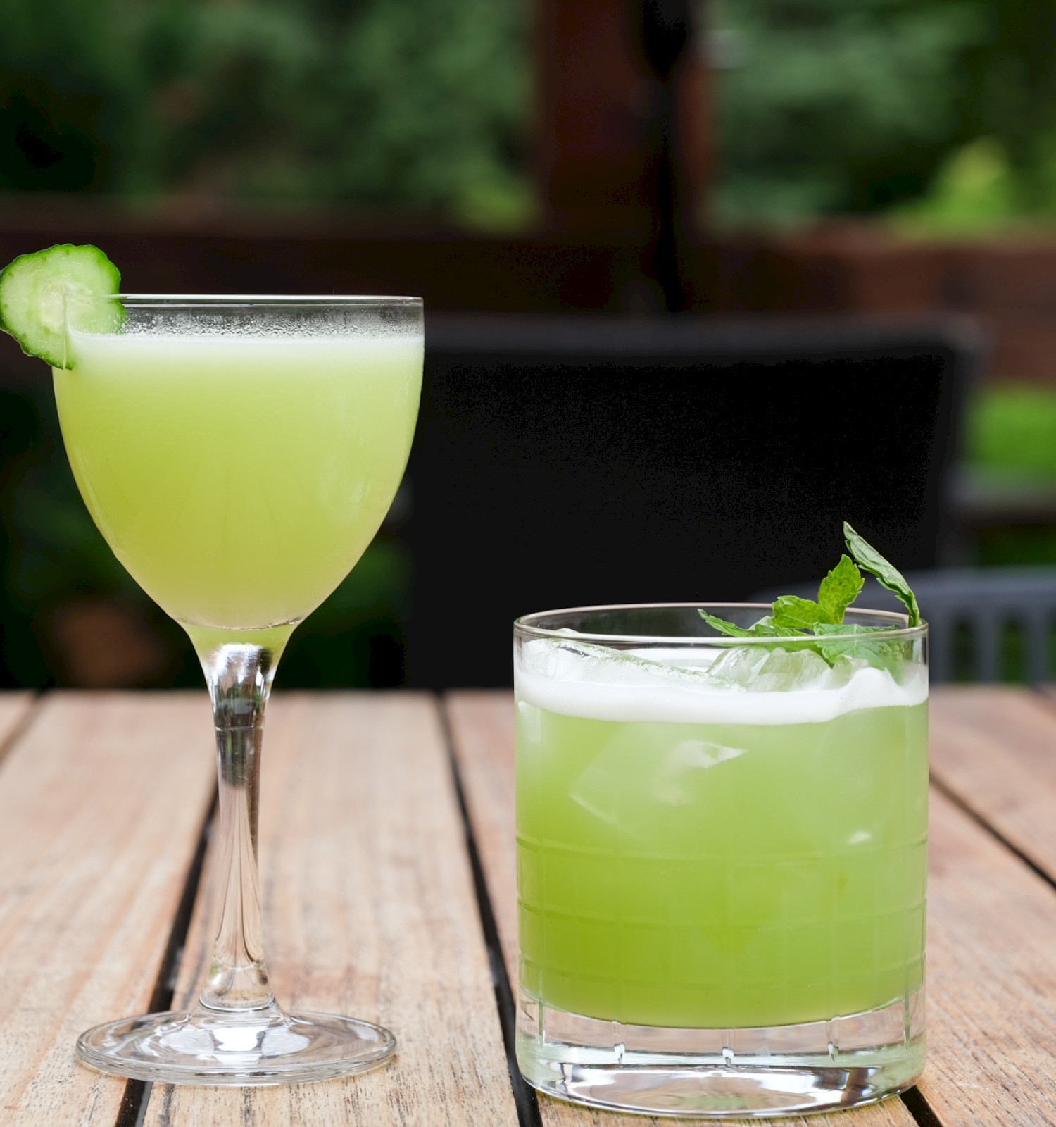 Two green cocktails with mint leaves and cucumber garnishes sit on a wooden table outdoors.