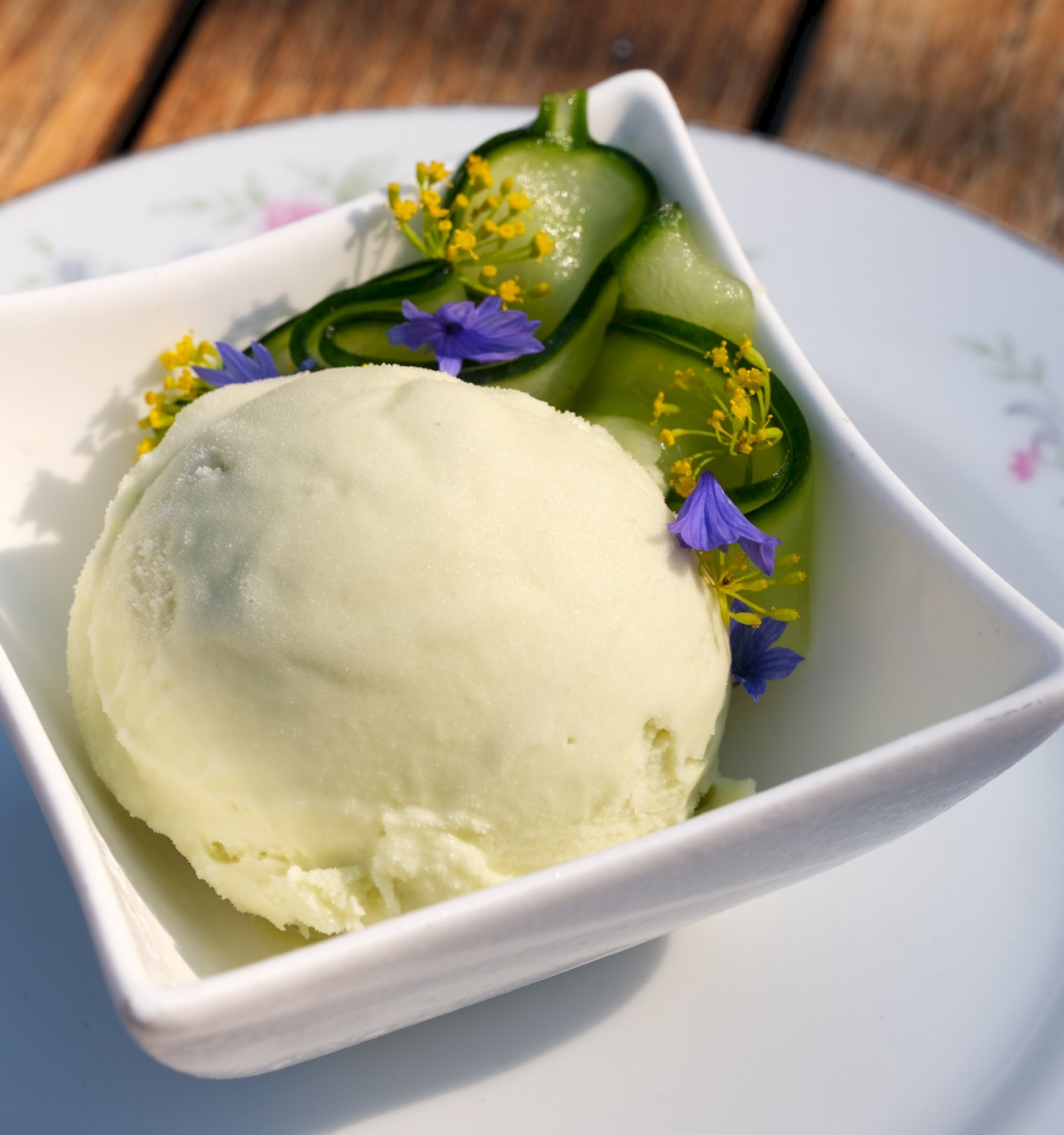 A bowl of vanilla ice cream with cucumber slices, yellow and purple edible flowers on a floral patterned plate outdoors.