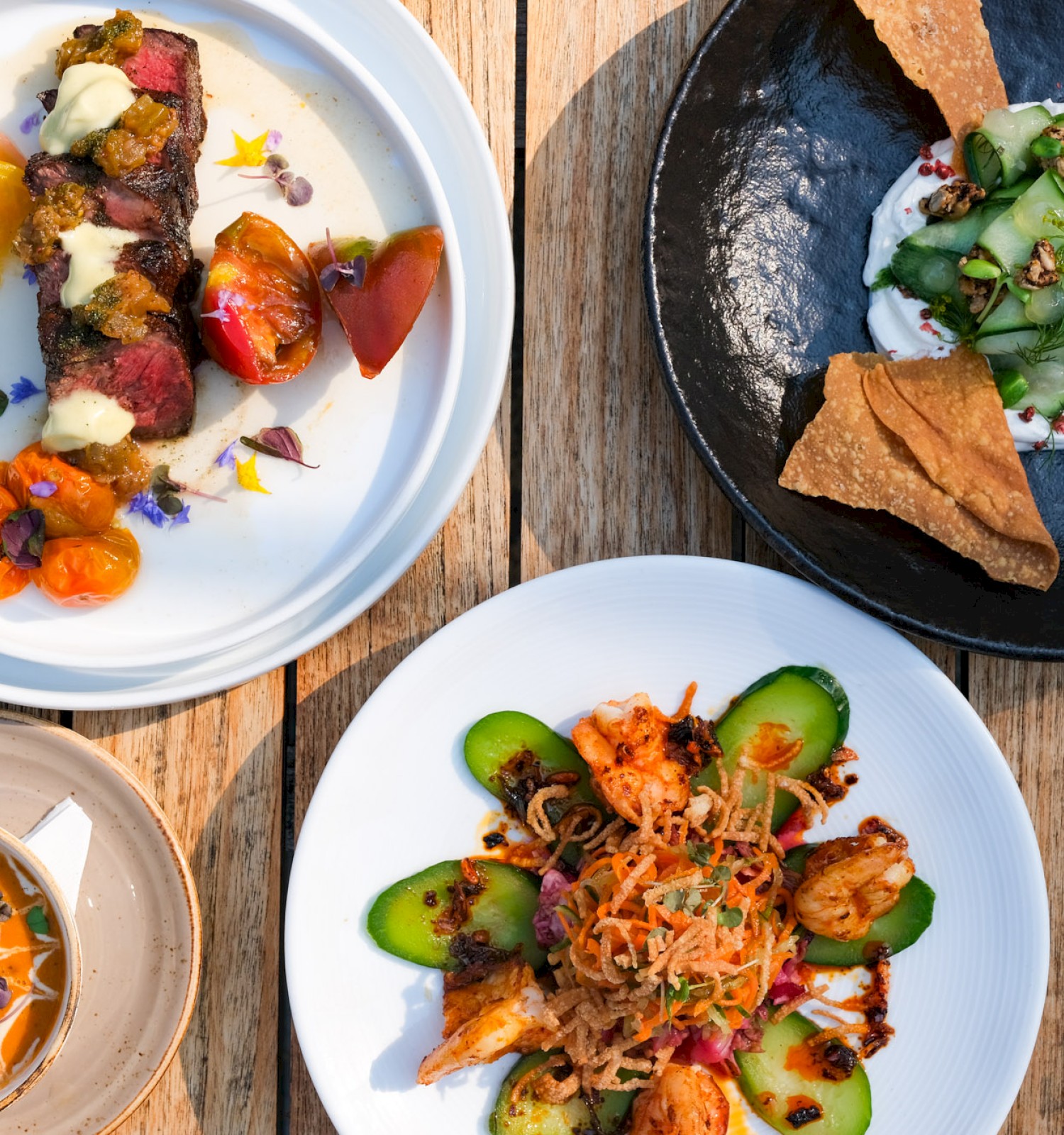 This image shows a variety of colorful, gourmet dishes arranged on a rustic wooden table, including a steak with tomatoes, a vegetable plate, a soup, and a salad.