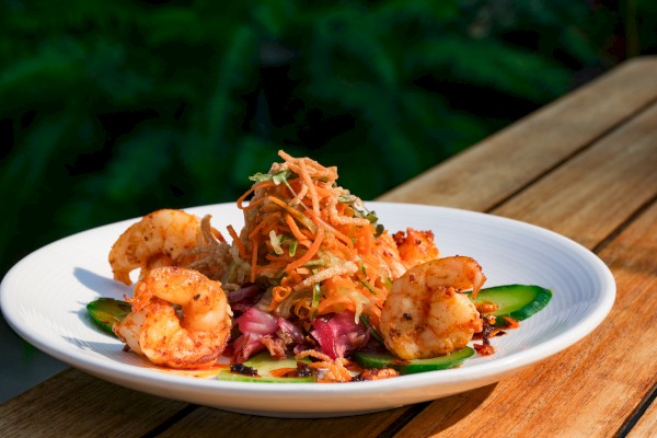 A plate of grilled shrimp with colorful mixed salad and vegetable garnishes on a wooden table, with a blurred green background.