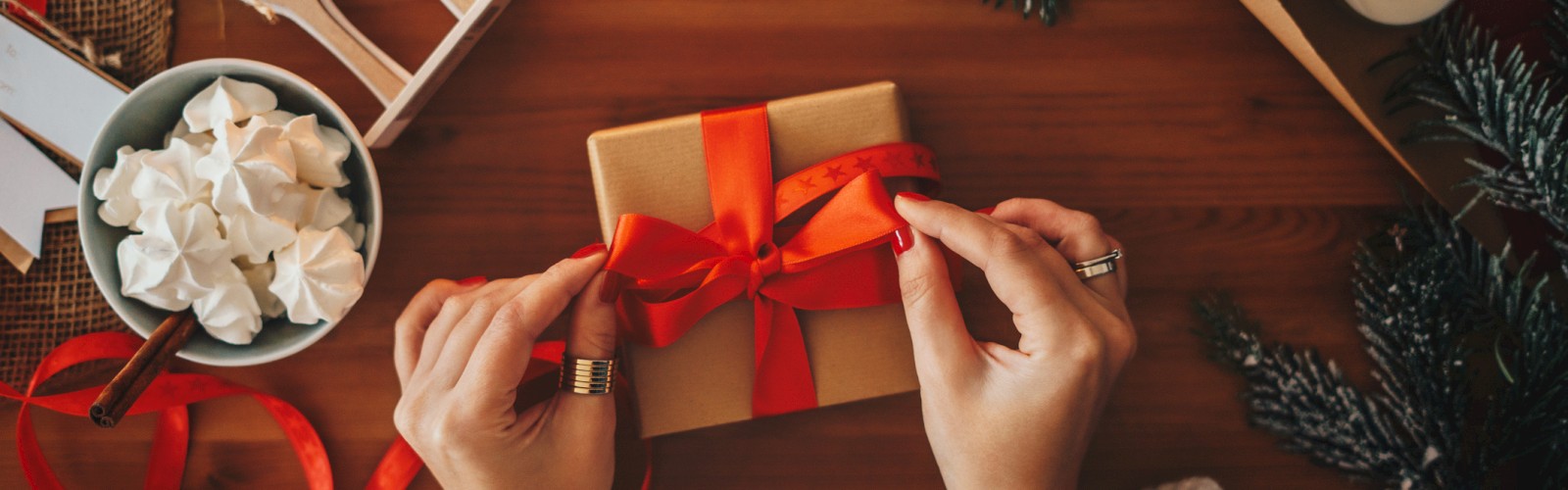 Person hands tying a small gift box with a red ribbon on a festive wooden table, surrounded by candles, pine branches, and treats, cozy holiday scene.