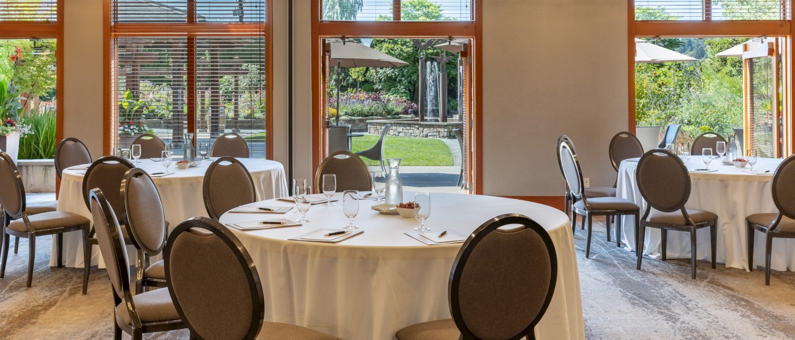 An elegantly set indoor dining area with round tables, white tablecloths, and chairs, overlooking a lush garden with umbrellas.