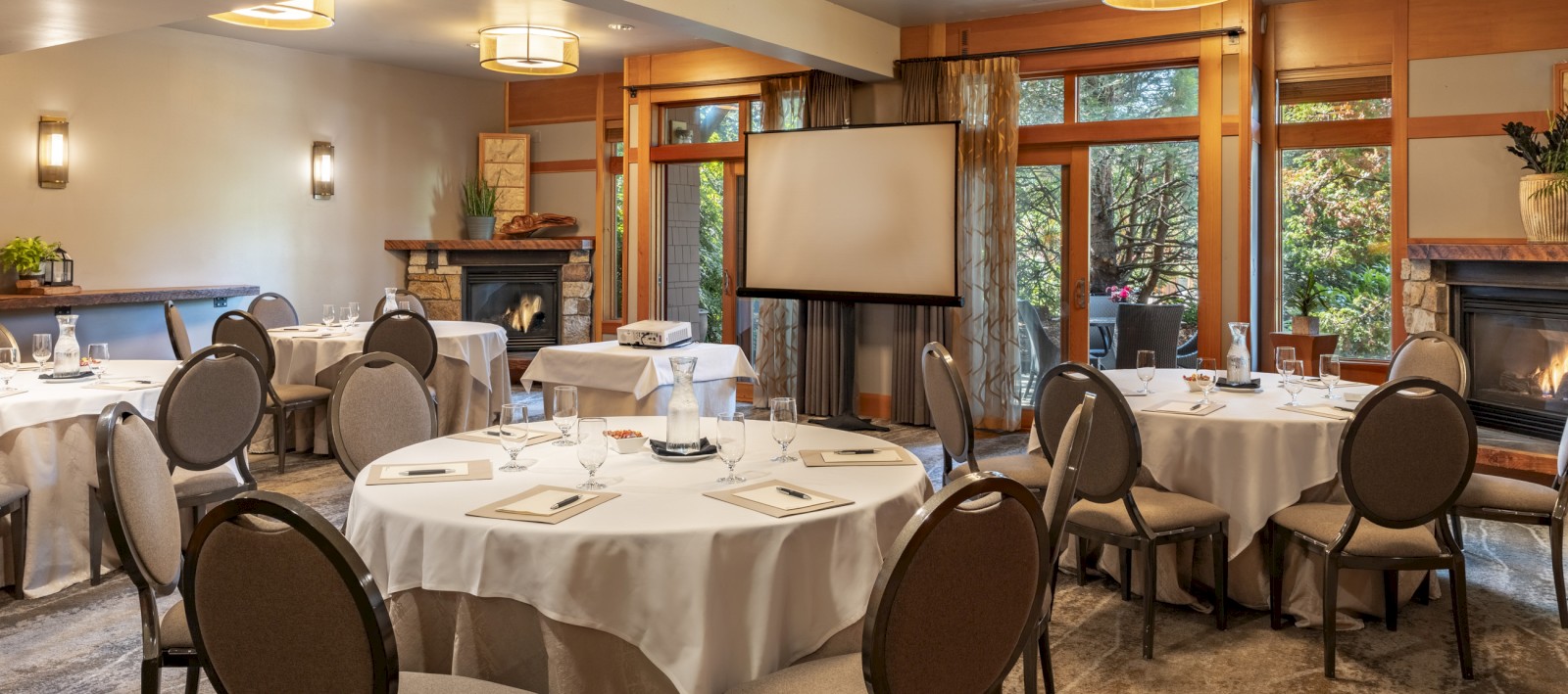A cozy, well-lit dining room set for a meeting or event, with round tables, chairs, a projector, and large windows showing greenery.