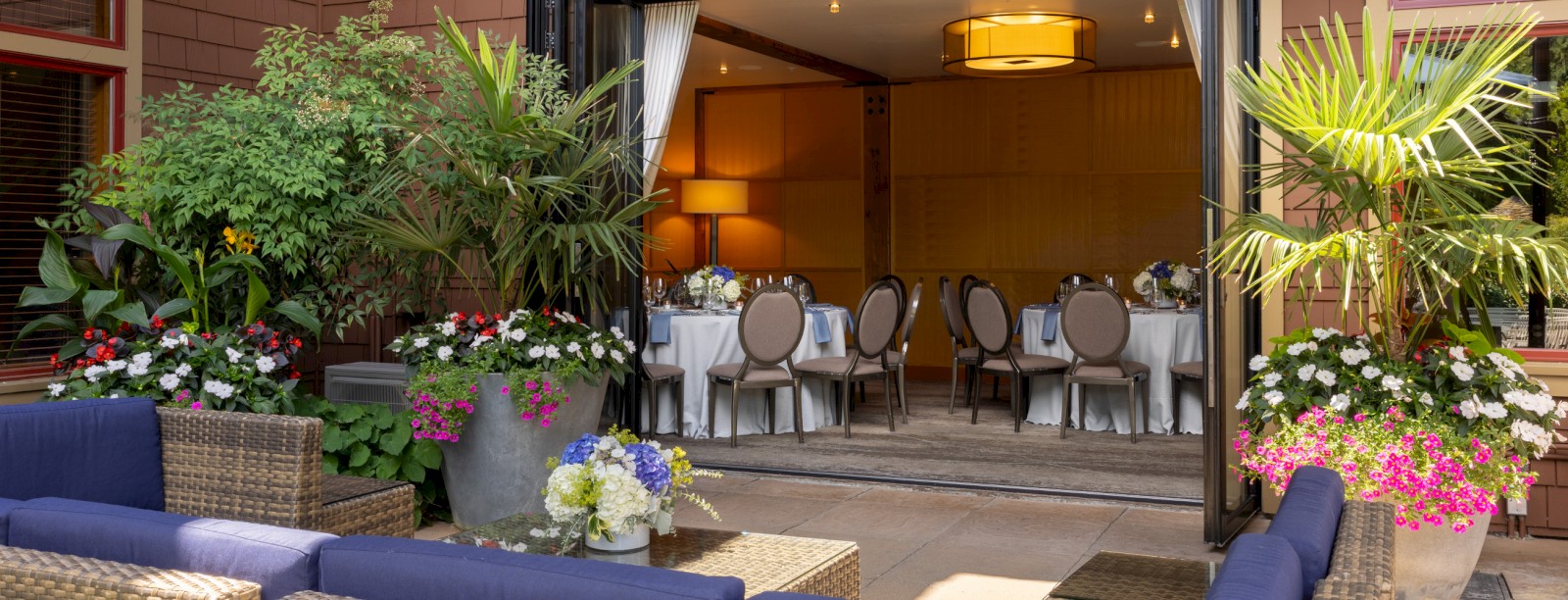 The image shows an outdoor dining area with flowering plants and a view of a cozy indoor restaurant setting.