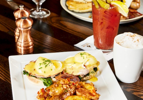 A plate with eggs Benedict, crispy potatoes, a Bloody Mary cocktail with garnishes, and a frothy cappuccino served on a wooden table.