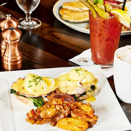 A plate with eggs Benedict, crispy potatoes, a Bloody Mary cocktail with garnishes, and a frothy cappuccino served on a wooden table.