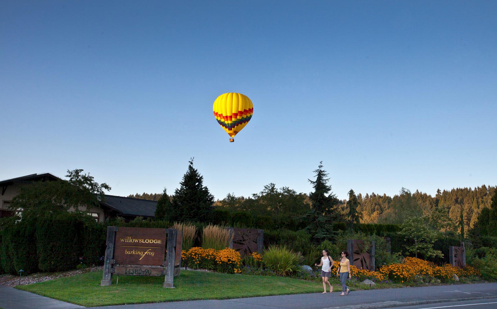 A hot air balloon flies over a scenic area with a building and gardens, a sign in front reads 