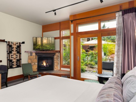 A cozy bedroom with a fireplace, a TV showing a forest scene, and large windows and doors opening to a garden area.