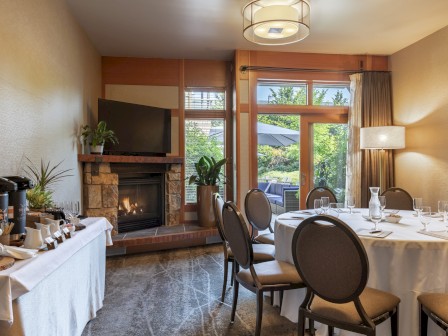 A cozy hotel room with a fireplace, a round dining table set for a meal, and outdoor seating visible through large windows.