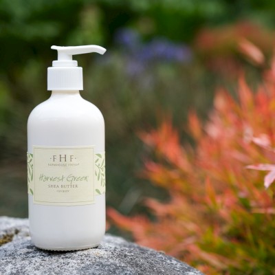 A white bottle of Farmhouse Fresh Harvest Green Shea Butter for body on a rock outdoors with colorful plants in the background.